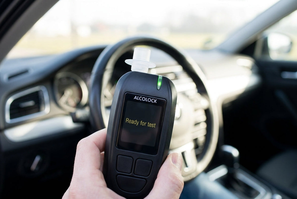 Driver seated in a car holding an ALCOLOCK AL-2500 ignition interlock device that reads Ready for test, before the start-up breath test